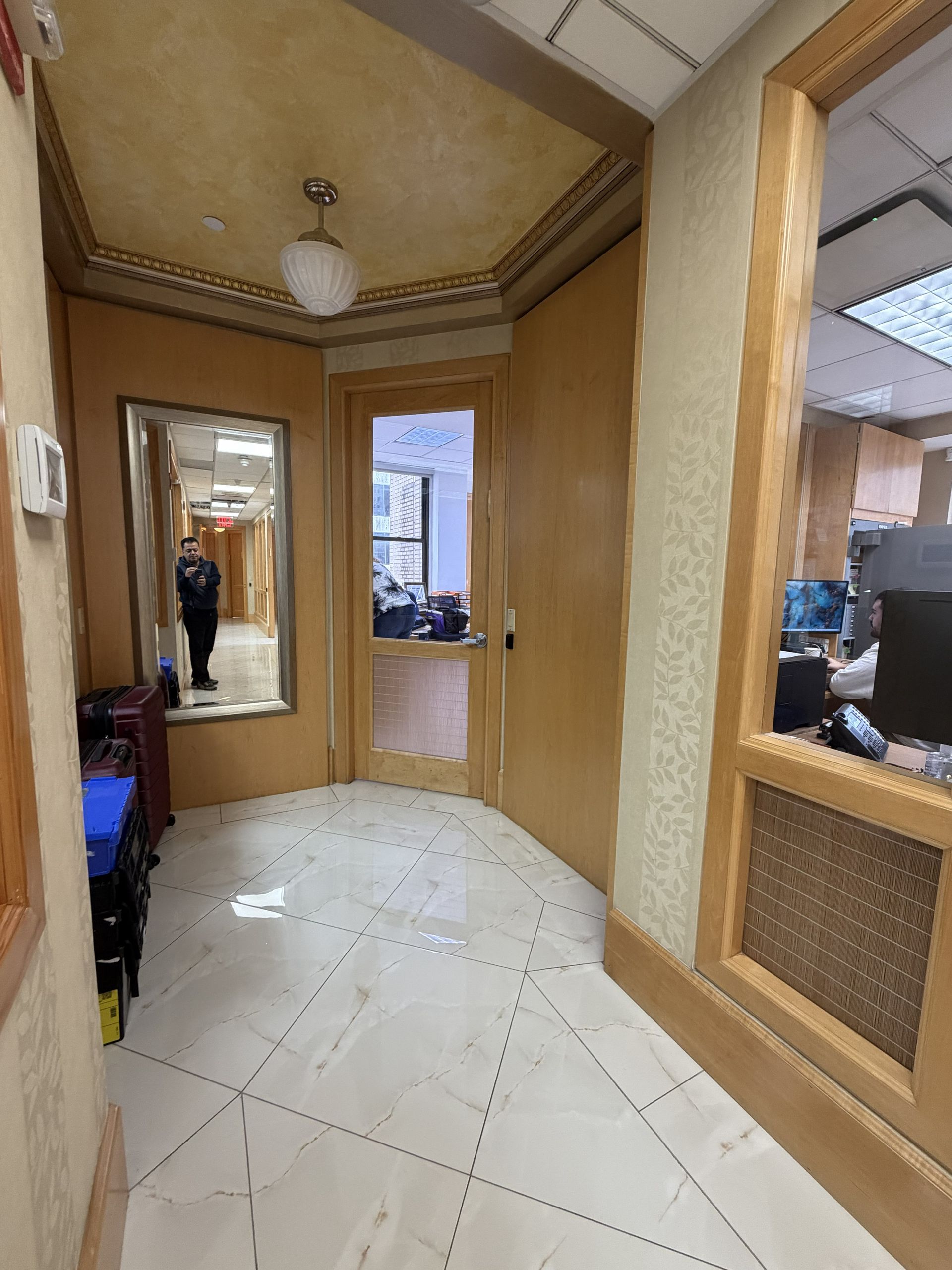 Narrow hallway with wood-paneled walls, white tiled floor, and a mirror reflecting a person near the entrance.