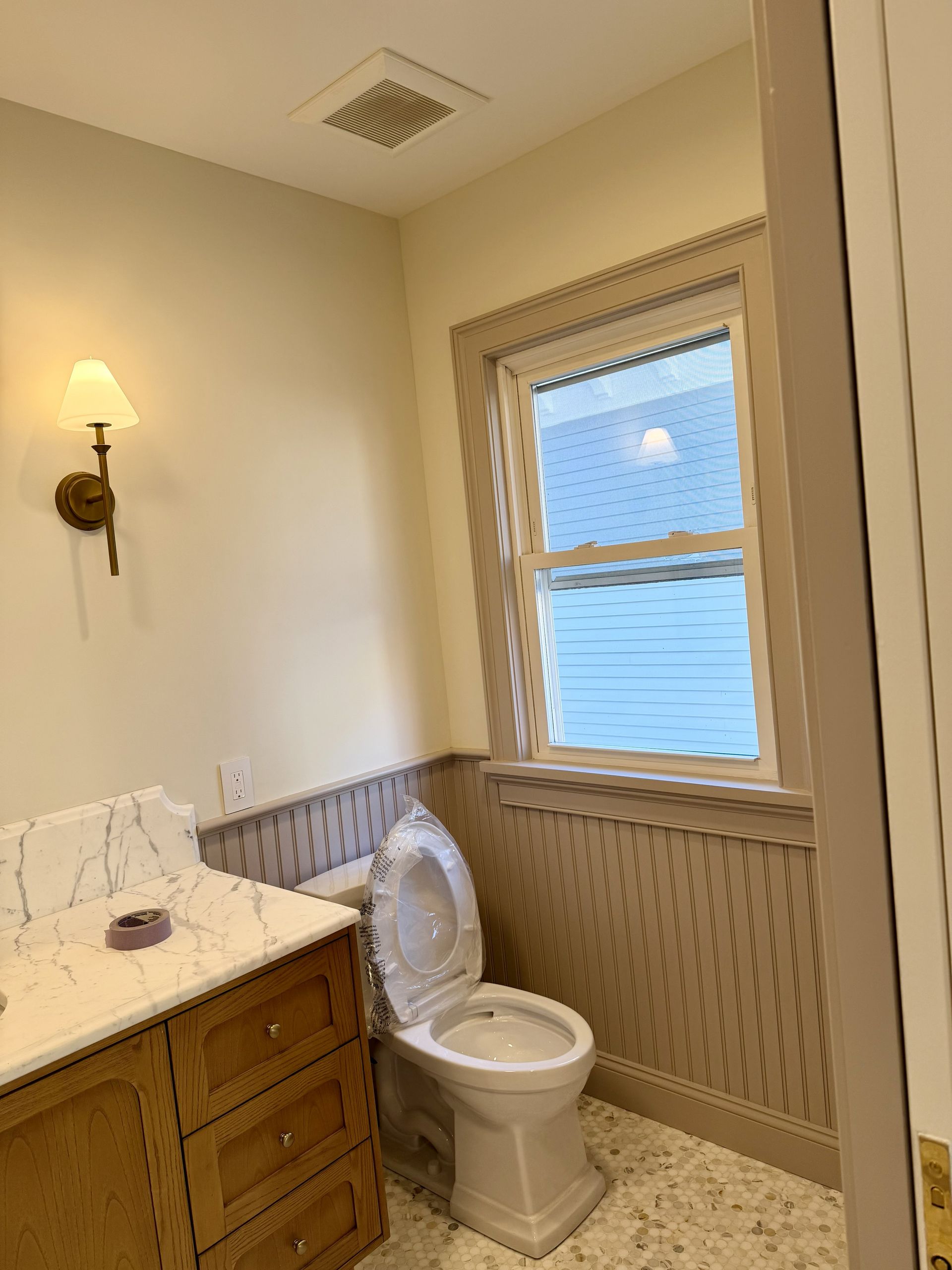 Small bathroom with toilet, wood vanity, wall sconce, and window with blinds.