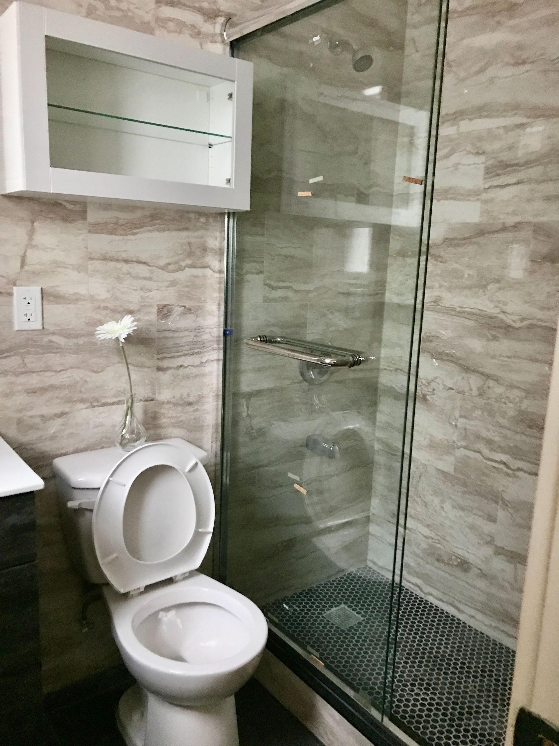 Small bathroom with toilet, glass shower, and wall-mounted cabinet in beige tile.