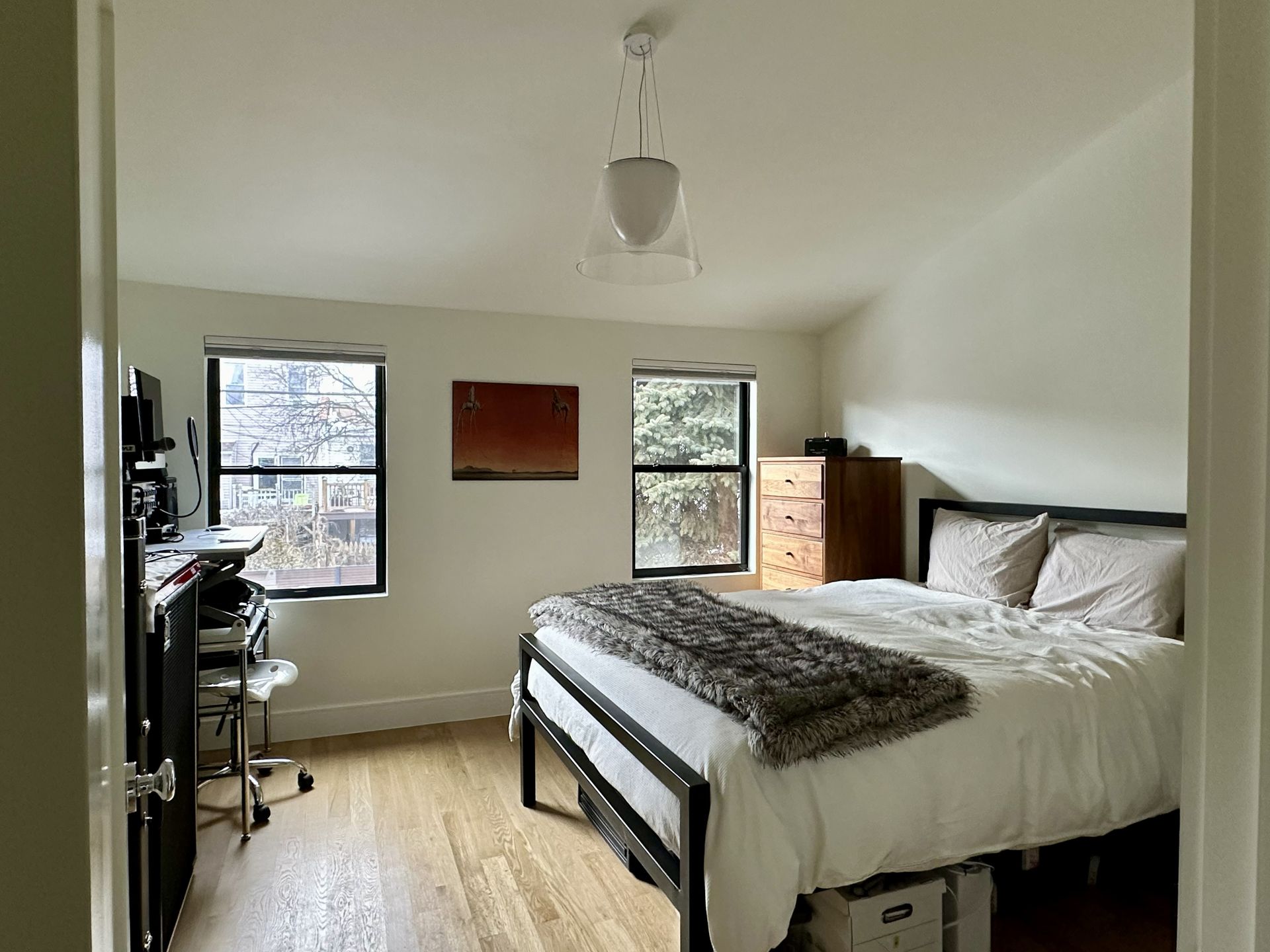 Bright bedroom with bed, white walls, two windows, and a small desk by the left window.