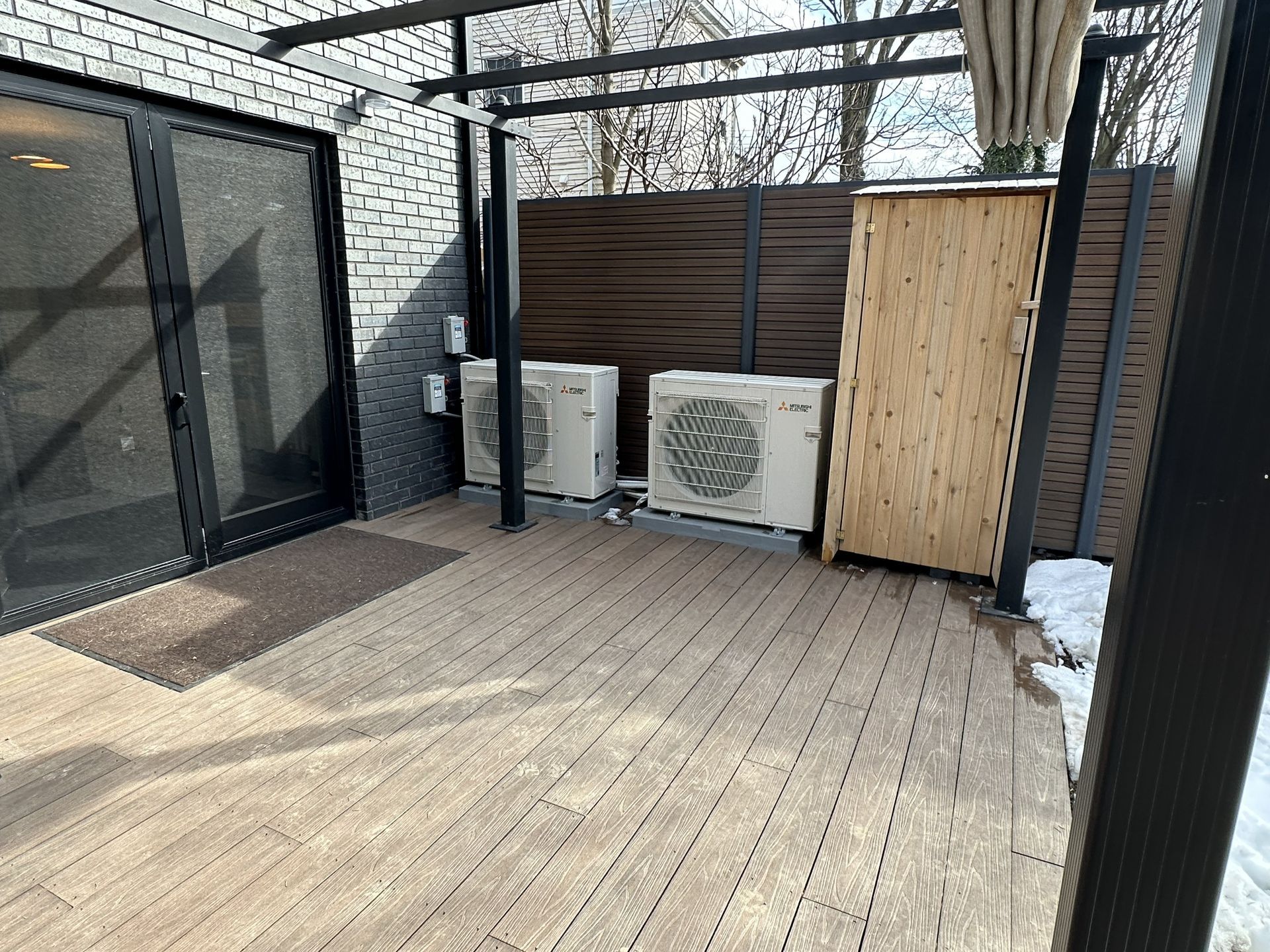 Small patio deck with a black-framed door, wooden fence, and two outdoor AC units under a pergola grid.