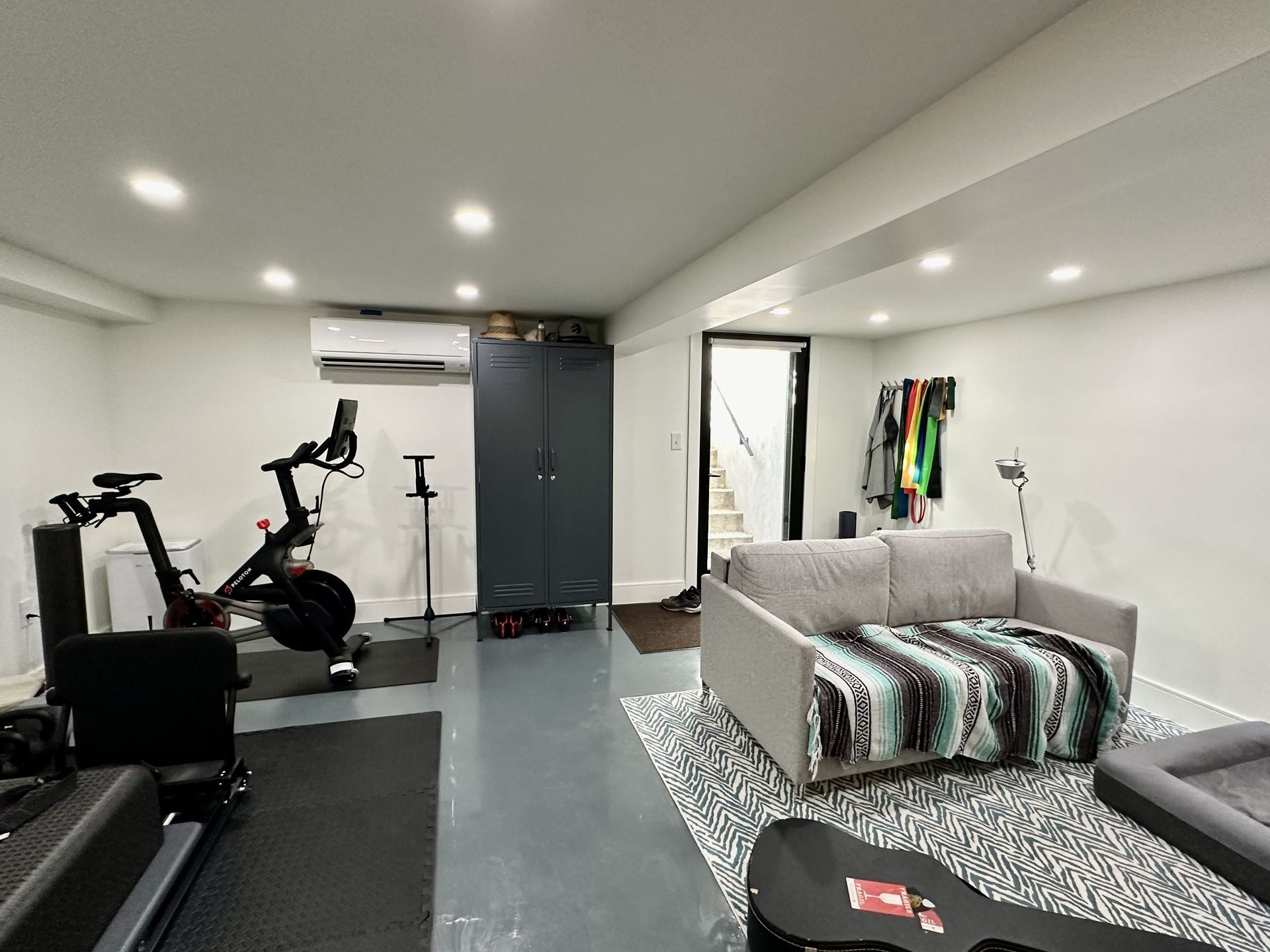 Home gym with treadmill, exercise bike, wall-mounted TV, gray cabinet, and a couch in a bright basement.