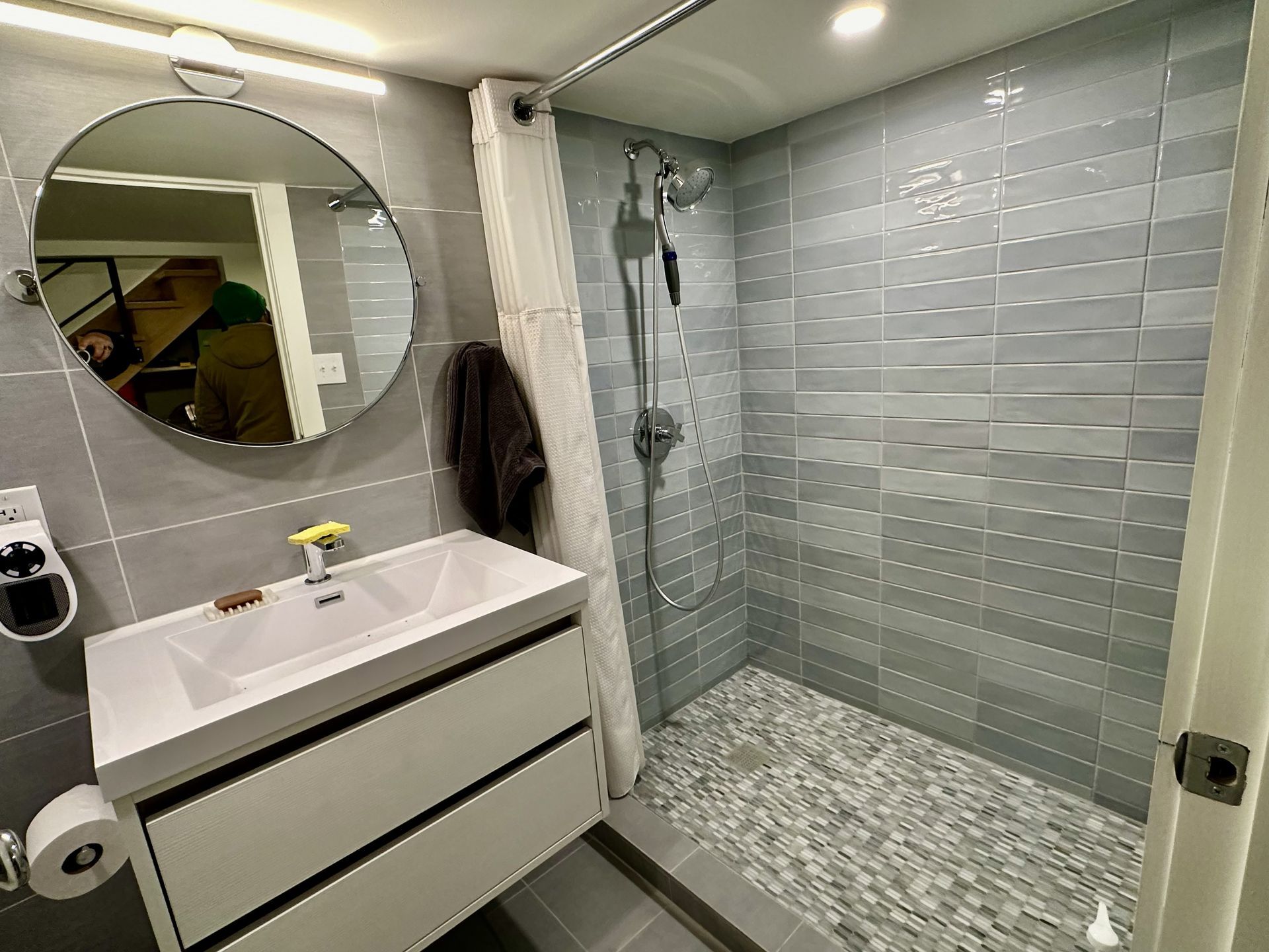 Modern bathroom with white vanity, round mirror, gray tiled shower, and frosted glass enclosure