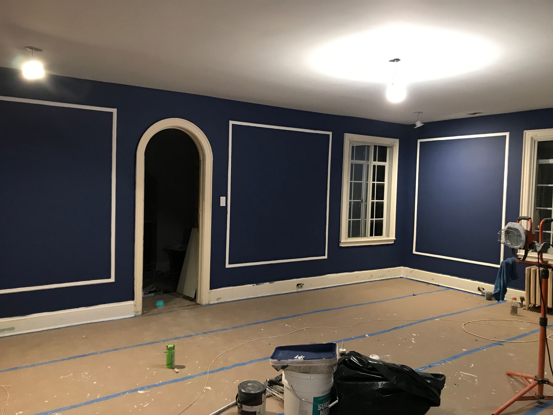 Blue room under renovation with taped wall trim, arched doorway, window, and painting supplies on the floor