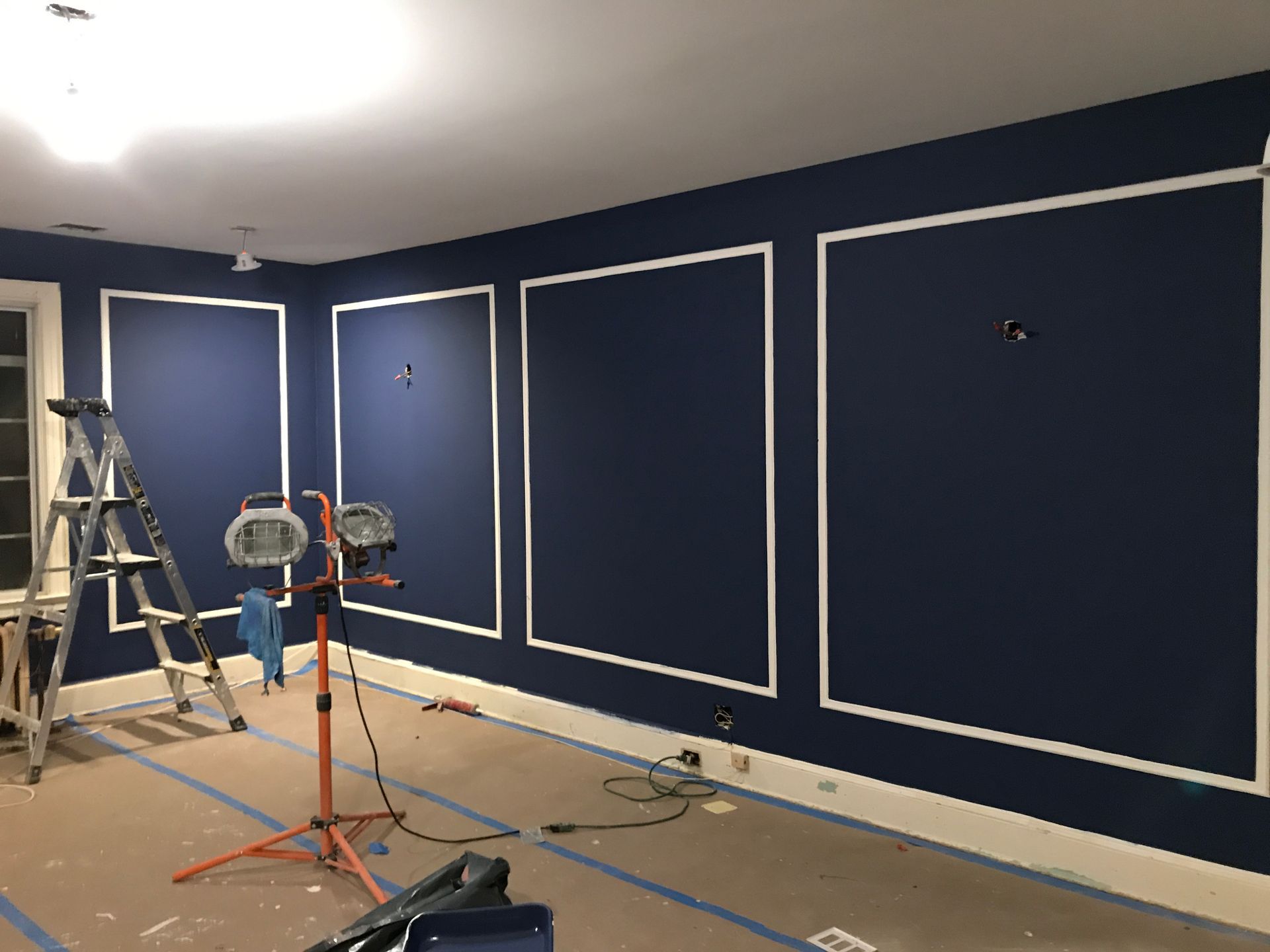 Blue room with white trim, ladders, and work lights during interior painting or renovation