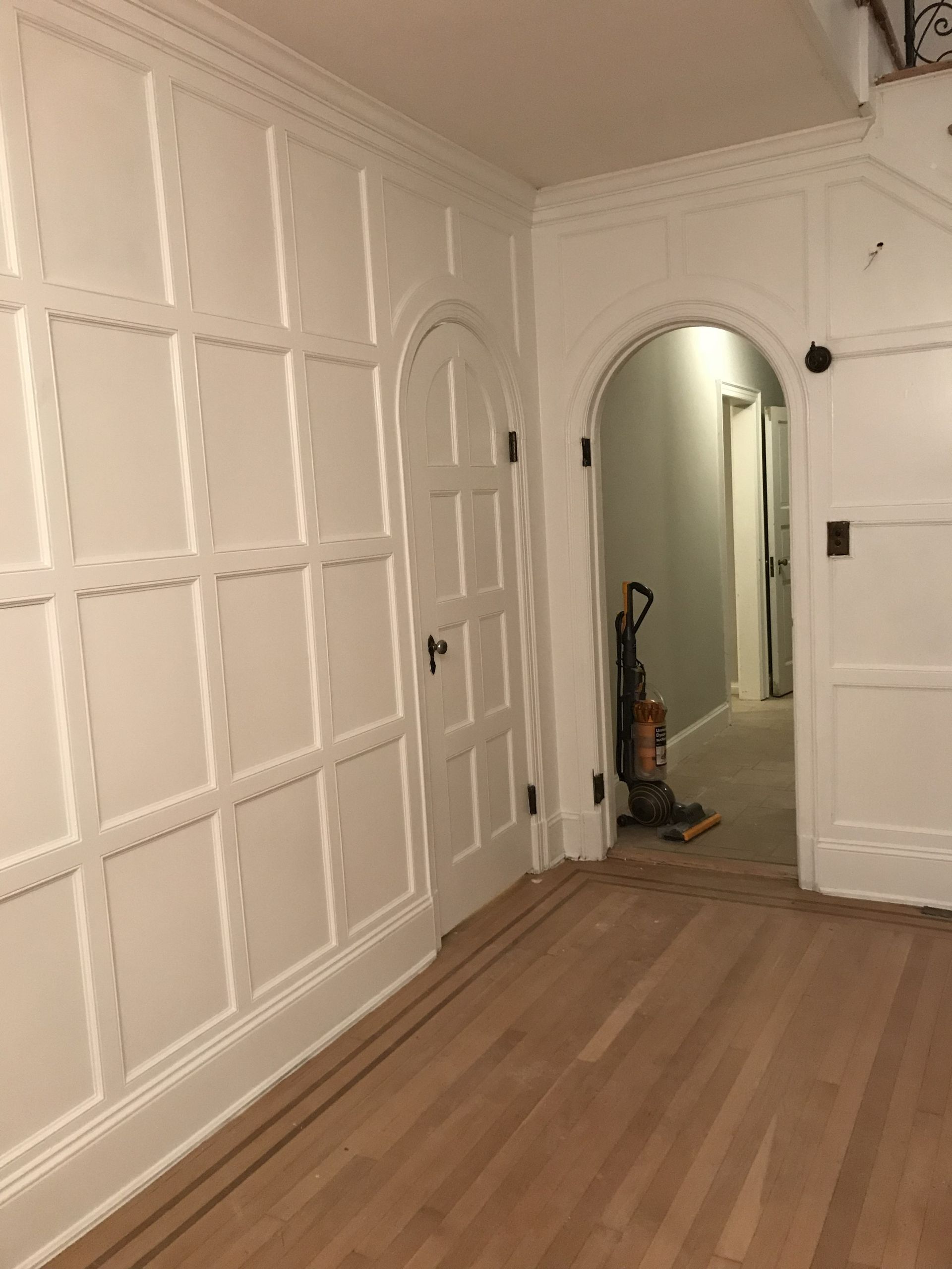 White paneled hallway with hardwood floor and arched doorway, showing a vacuum in the background
