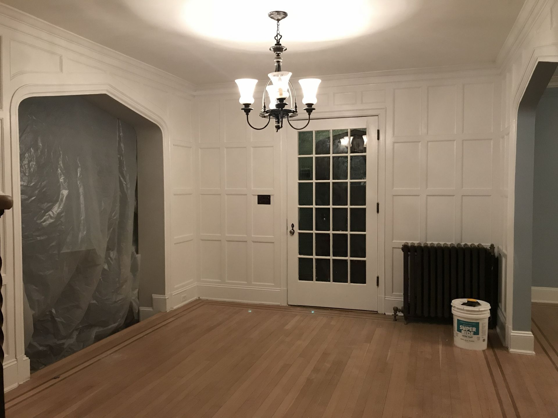 Empty room with paneled white walls, hardwood floor, chandelier, French door, radiator, and a paint bucket.
