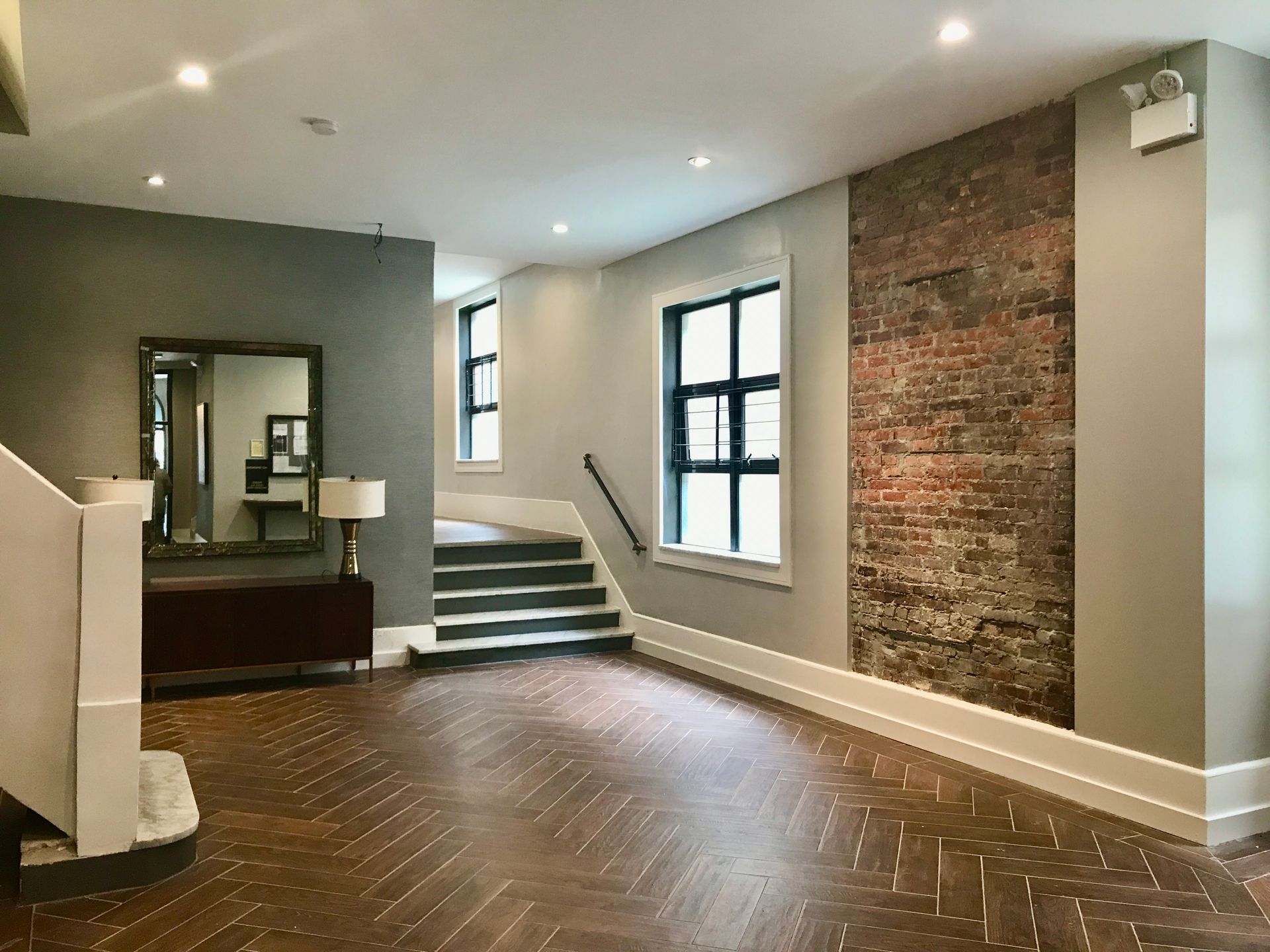 Empty modern room with exposed brick wall, dark wood floor, windows, and a small staircase.