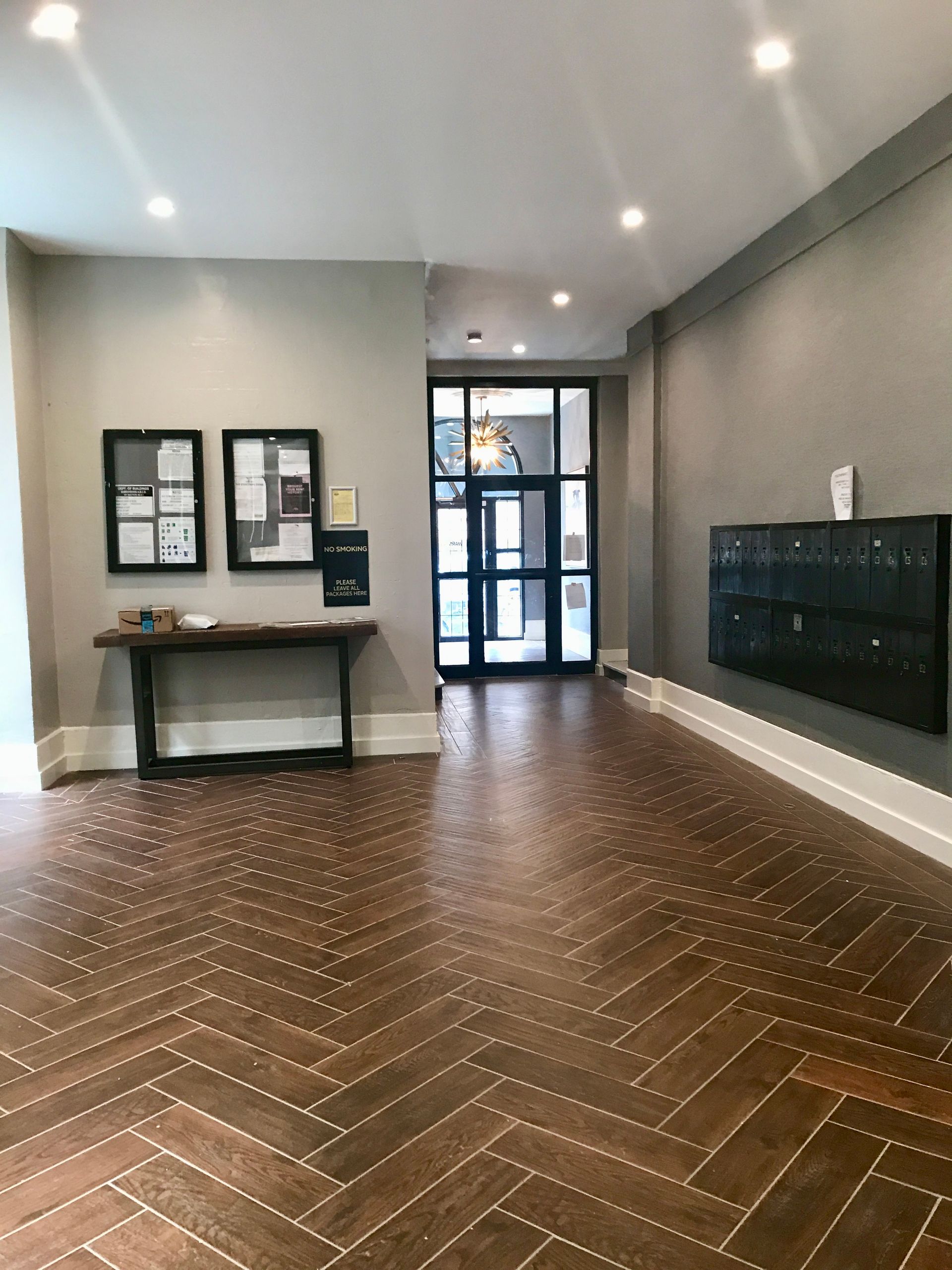 Empty lobby with herringbone wood floor, gray walls, wall-mounted display case, and glass entrance doors.