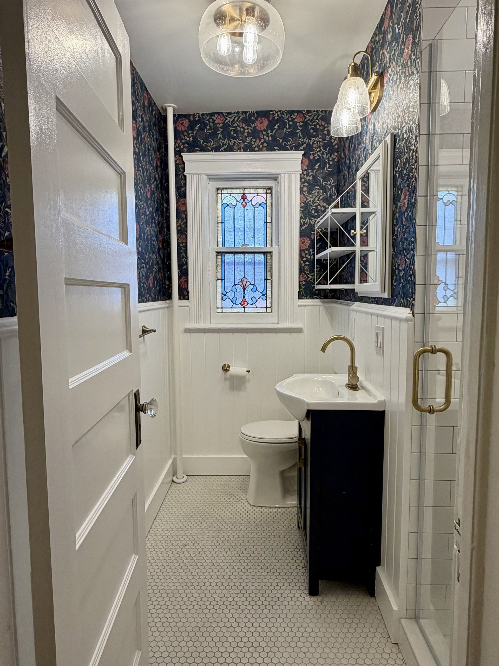 Narrow white bathroom with patterned wallpaper, pedestal sink, toilet, window, and glass shower door.