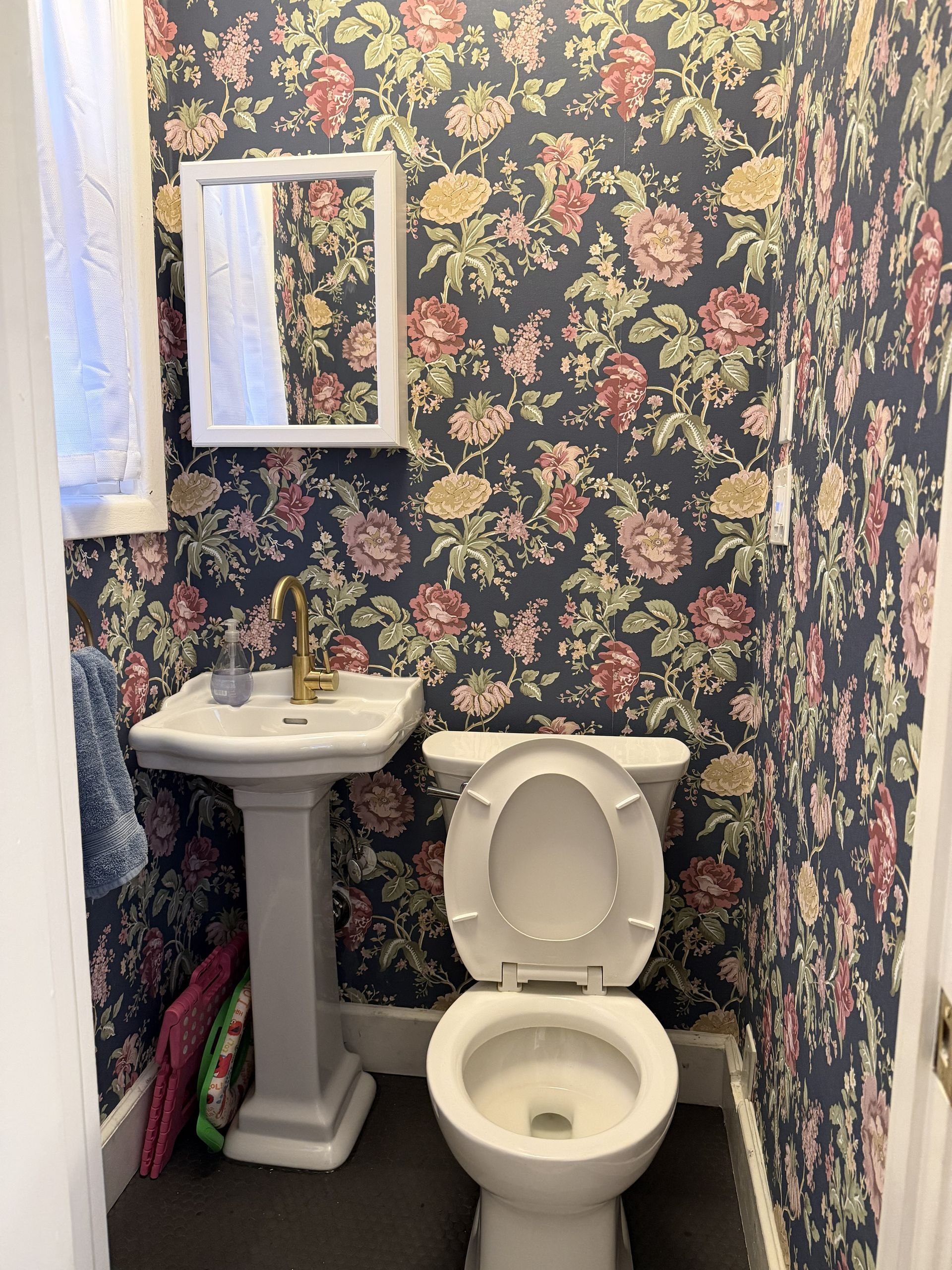 Small bathroom with floral wallpaper, pedestal sink, mirror, and open toilet seat.