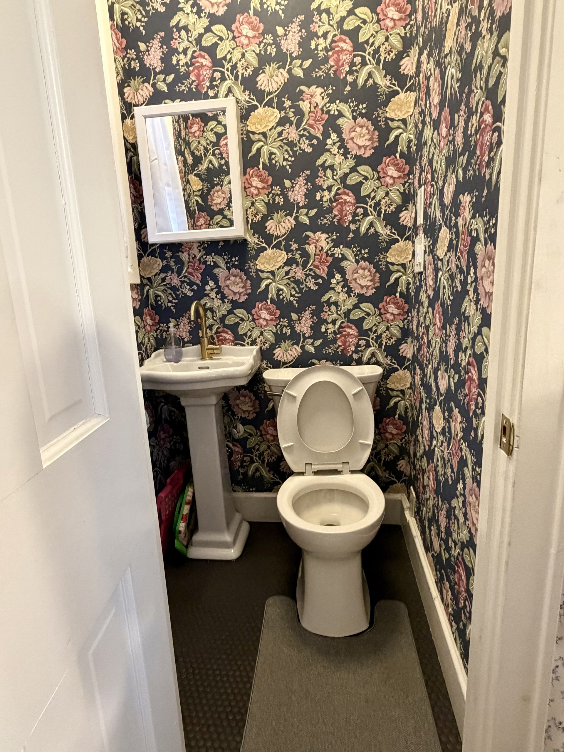 Small bathroom with floral wallpaper, pedestal sink, mirror, and open toilet seen through a doorway.
