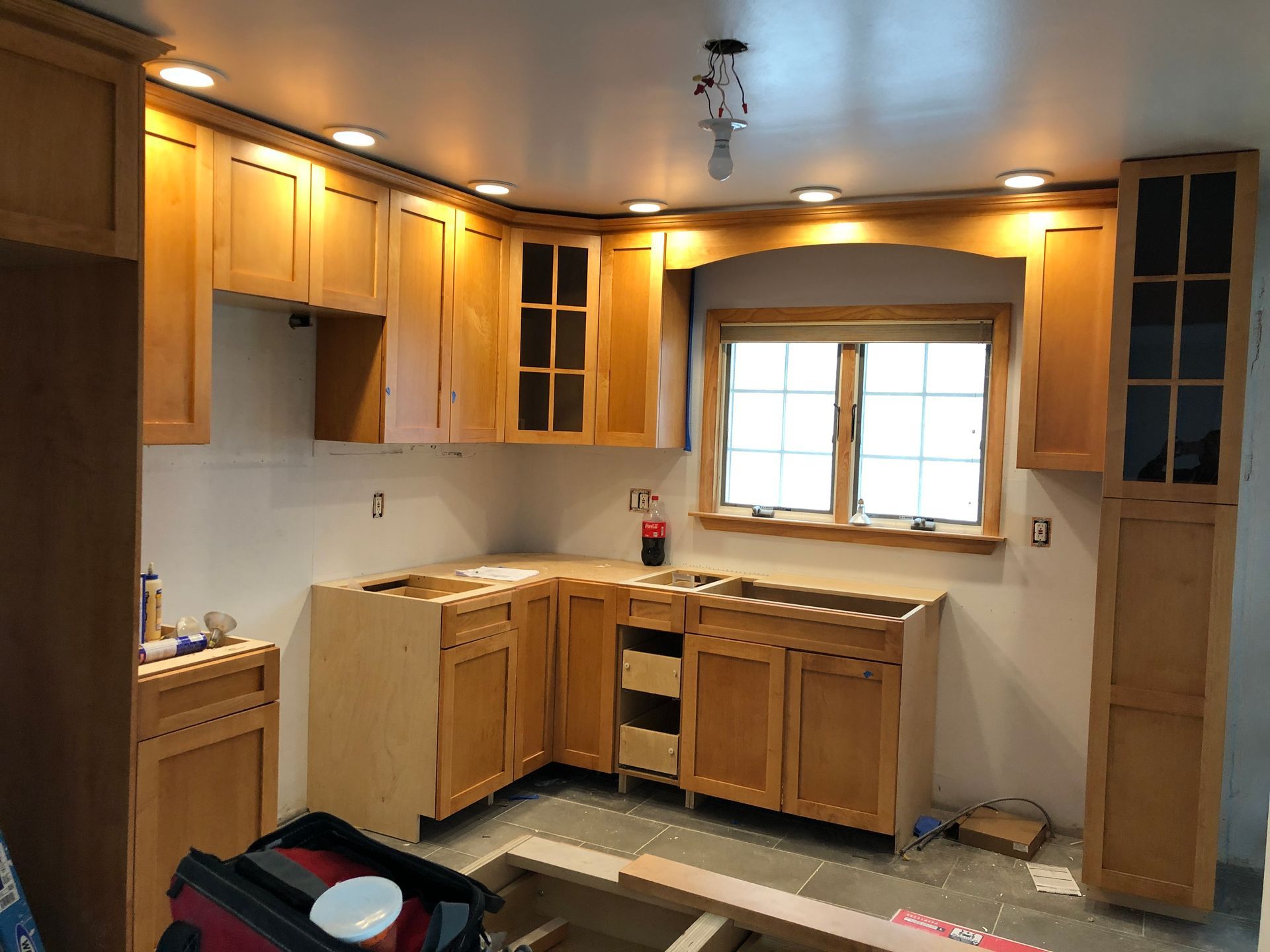 Unfinished kitchen with light wood cabinets, countertop, window, and tools on the floor.