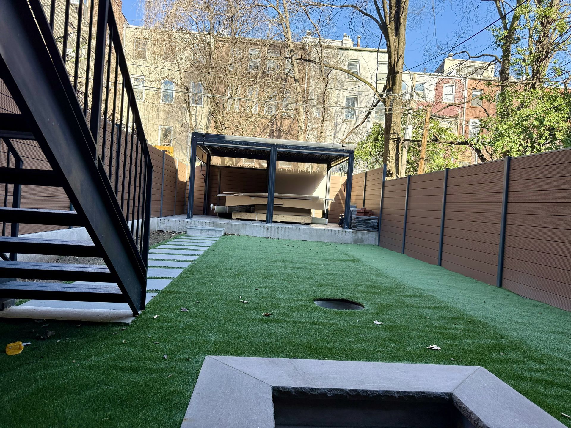 Narrow backyard with artificial turf, wooden fence, black stairs, and a covered patio in the background