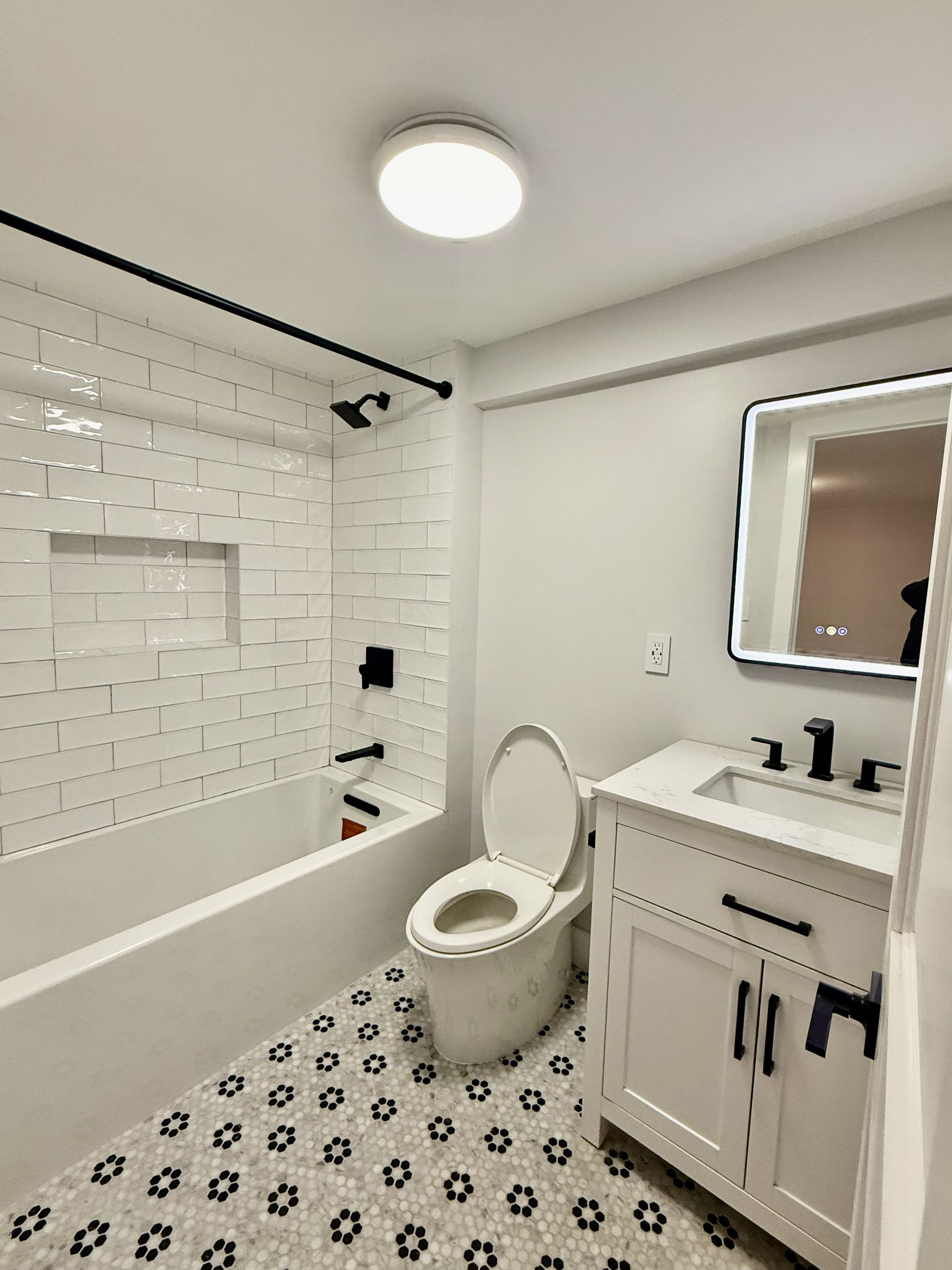Bright white bathroom with shower, toilet, vanity, and round illuminated mirror on patterned tile floor