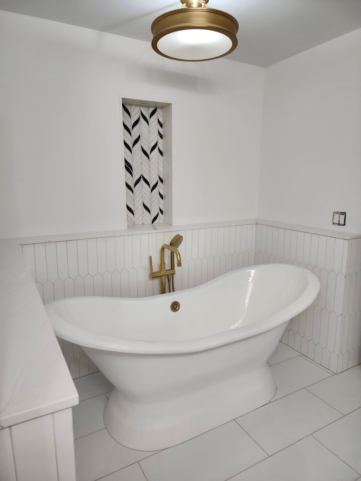 White bathroom with freestanding tub, gold fixtures, tiled floor and walls, and a narrow decorative wall niche.