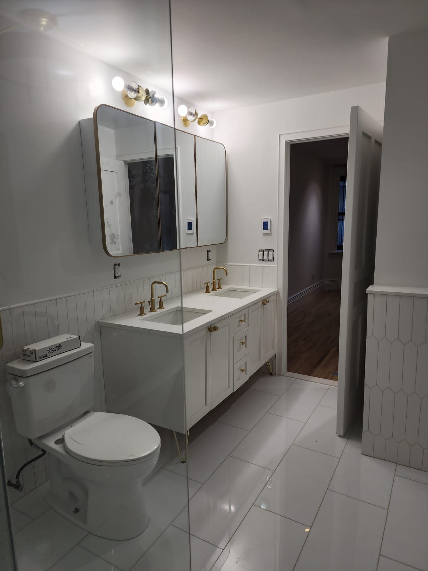 Bright white bathroom with double vanity, wall mirrors, toilet, and open door to hallway.