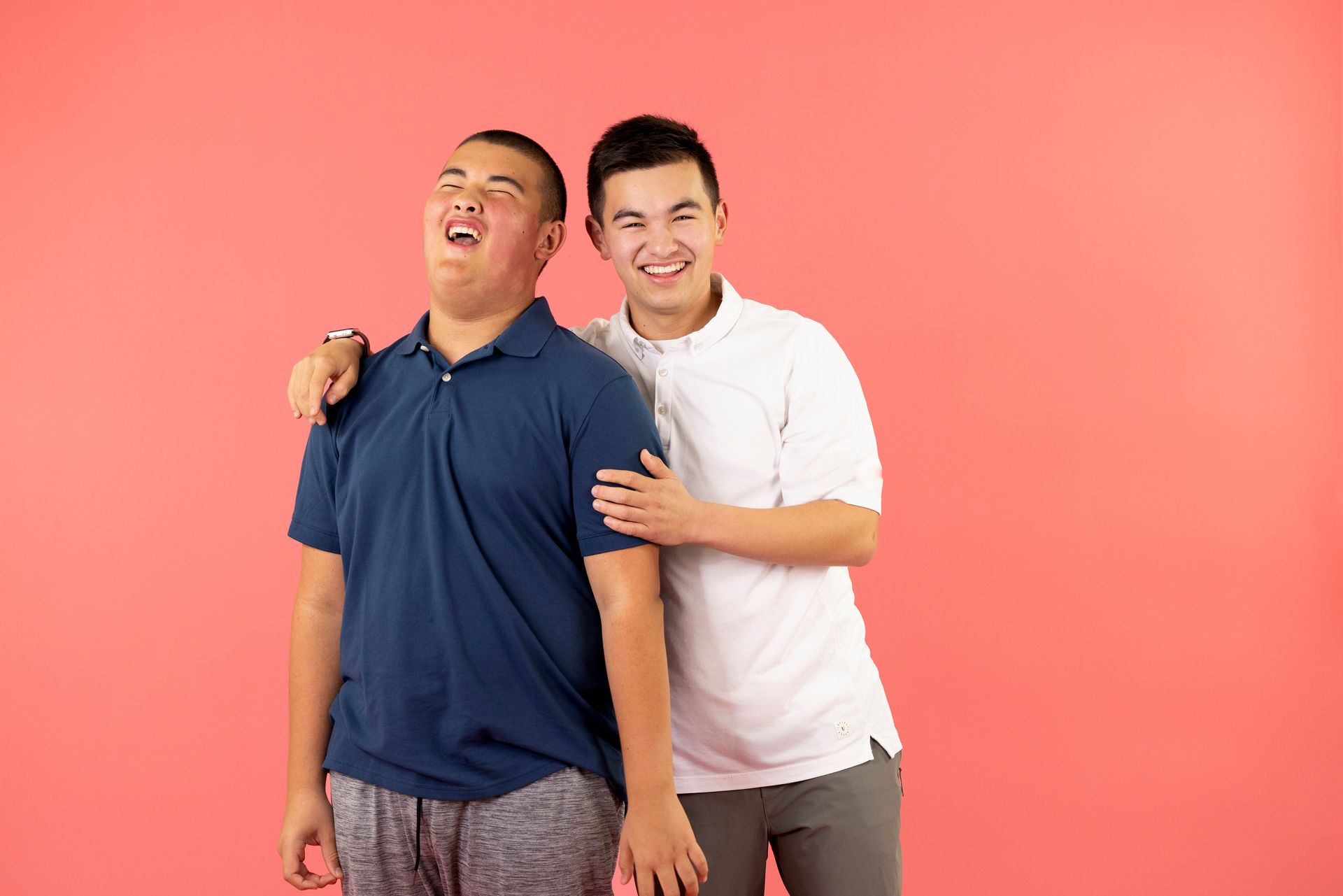 Two young men are standing next to each other on a pink background.