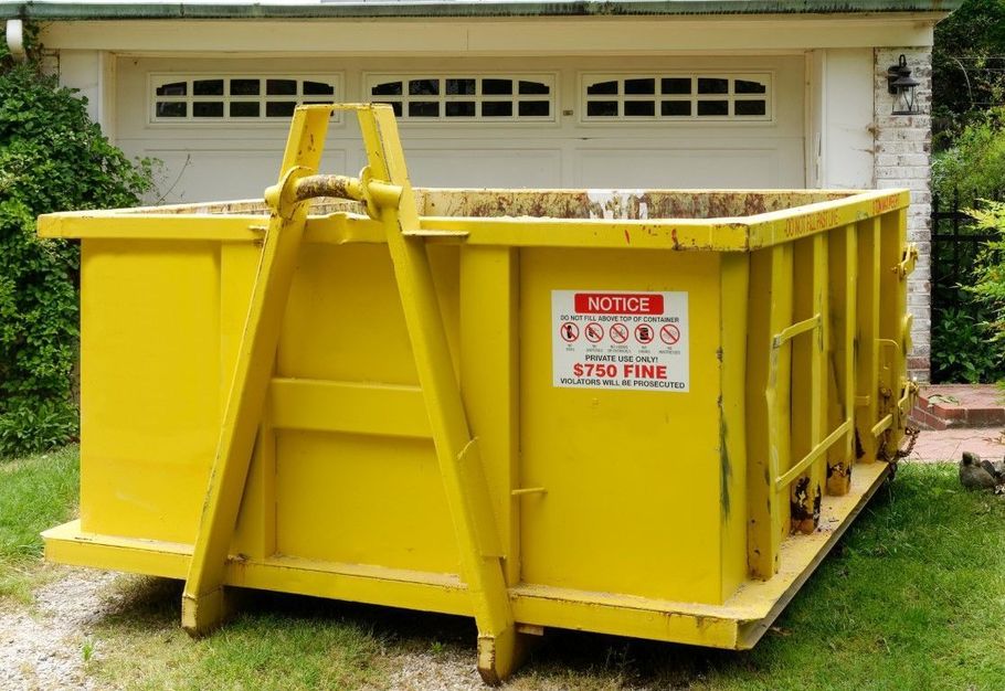 Yellow industrial dumpster/bin with warning sign, parked on grass in front of a building.