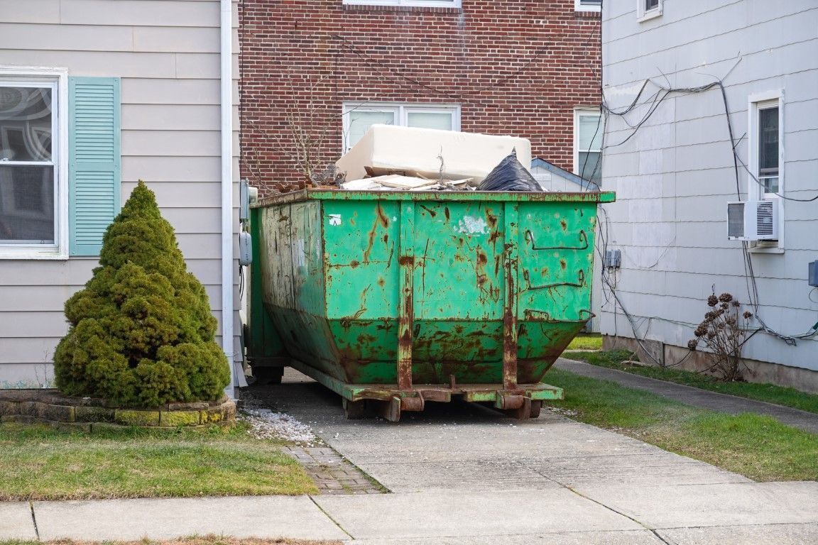 Green dumpster in a residential driveway beside a house and garage, with debris piled on top