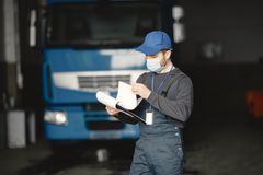 Trabalhador usando máscara facial e boné azul segura uma prancheta em frente a um caminhão azul estacionado em uma garagem.