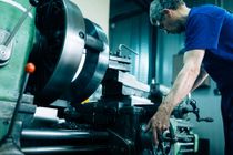 Uma pessoa de camisa azul operando um torno mecânico em uma oficina, concentrada na máquina.