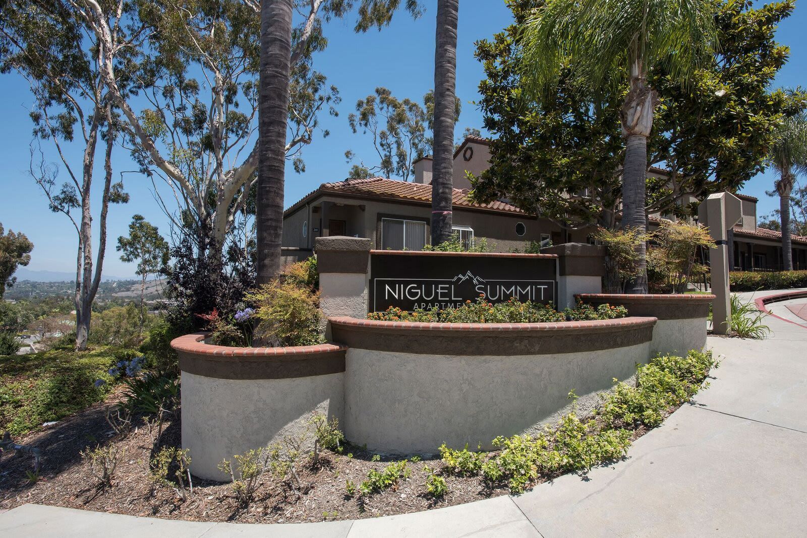Exterior entrance sign for Niguel Summit Apartments with palm trees and landscaped planters.