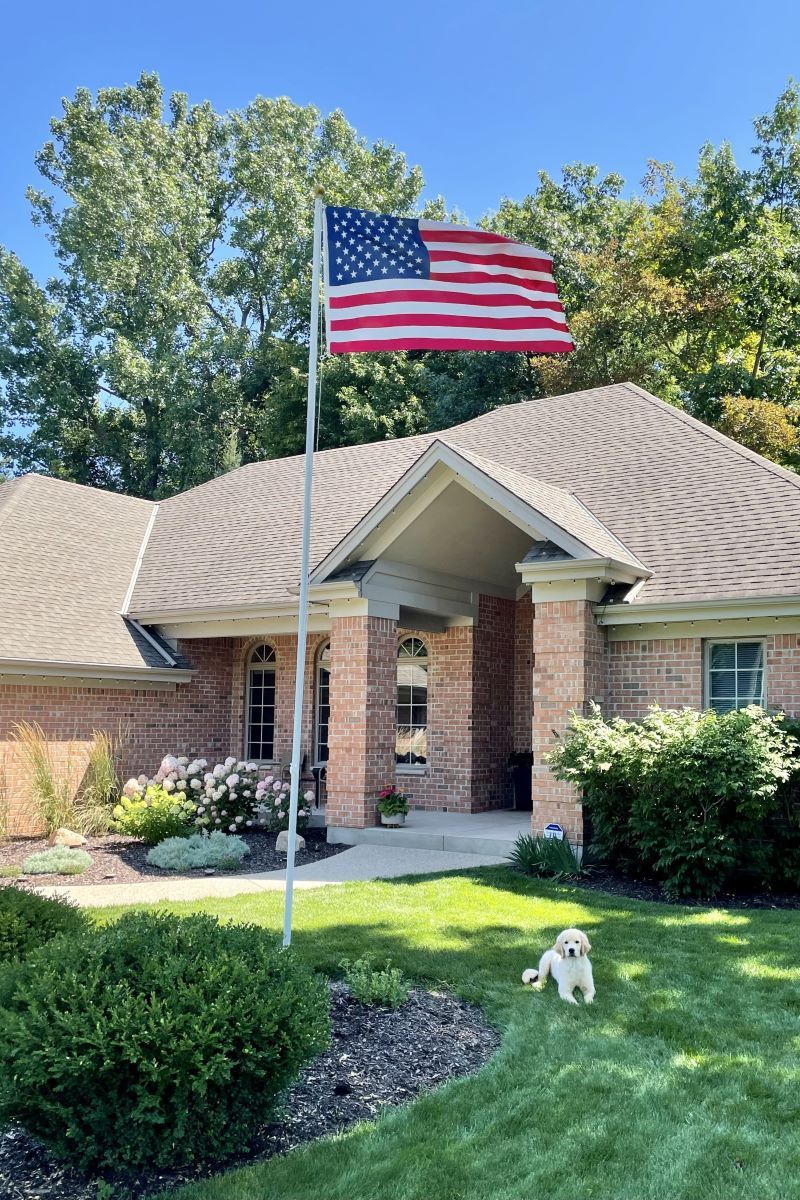 American flag on a pole in front of a brick house with a small dog on the lawn