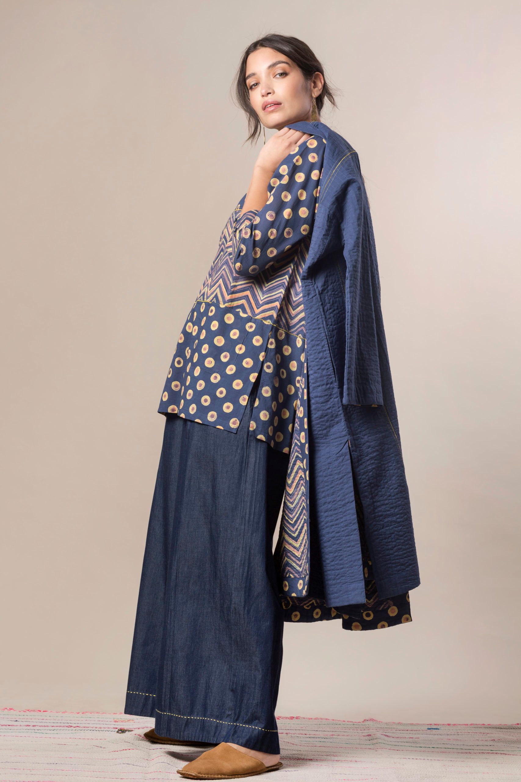 Donna in abito blu fantasia, pantaloni di jeans e cappotto, in posa su uno sfondo chiaro.