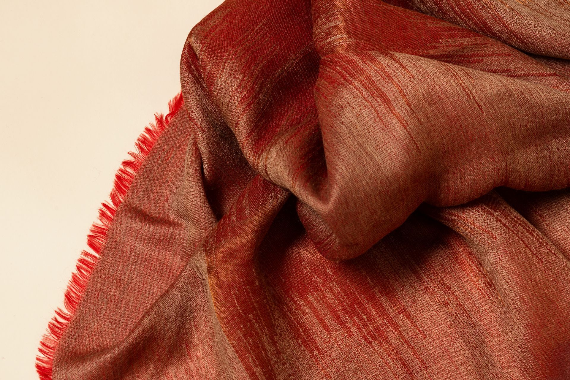 Sciarpa metallizzata rossa e oro con bordo sfrangiato, drappeggiata su uno sfondo color crema.