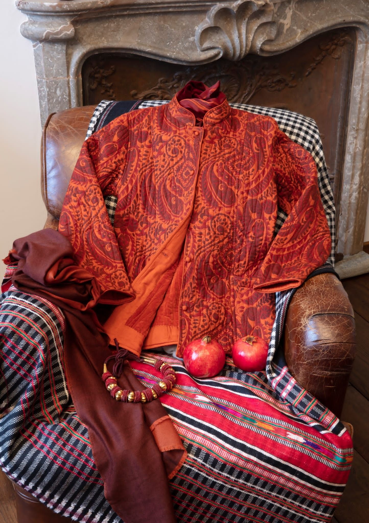 Giacca e sciarpa con fantasia rossa e arancione su una sedia in pelle, con mele e gioielli.