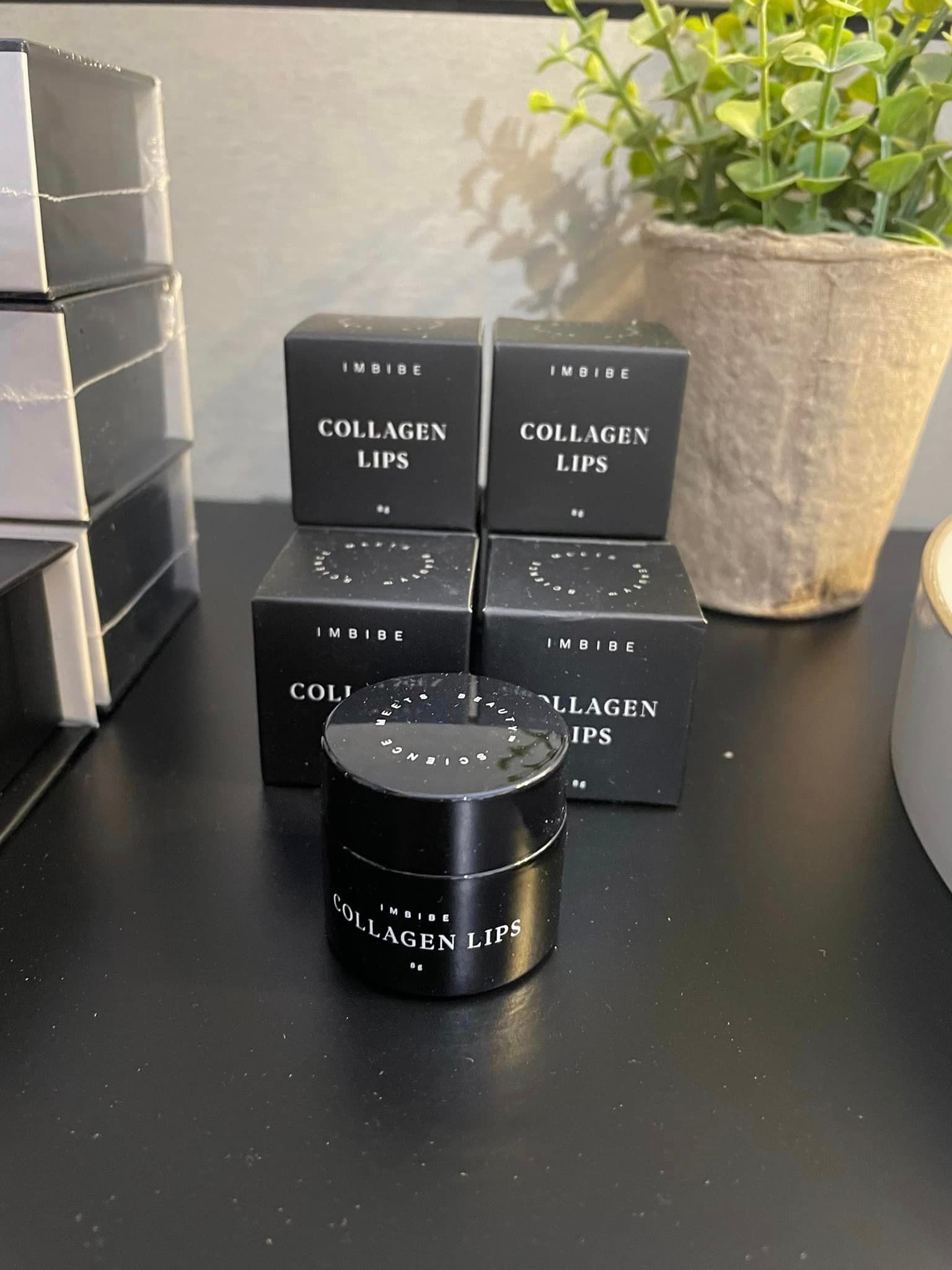 Black jars of collagen lip product on a black table with small boxes, a potted plant, and stacks of boxes.