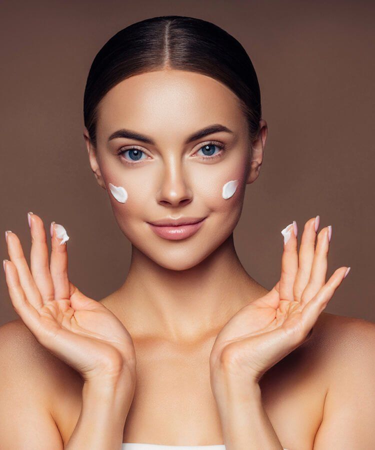 Woman with moisturizer on face and hands, smiling, brown background.