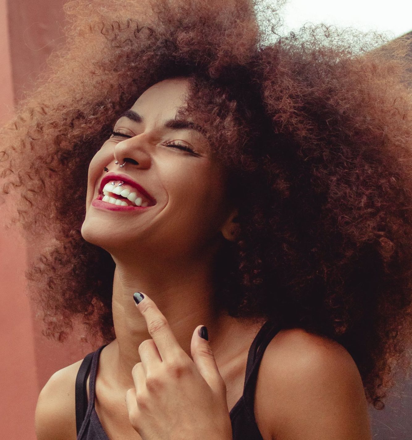 Woman with large curly hair laughs, pointing up, wearing a nose ring and black top.