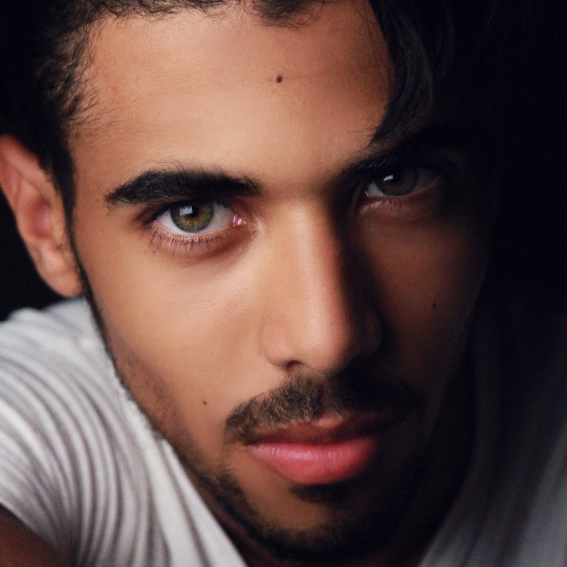 Man with dark hair and a beard, looking at the viewer. He wears a white shirt, with green eyes and a serious expression.