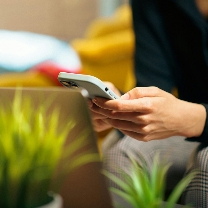 Hands holding a smartphone near a laptop, with blurred green plants and a yellow chair in the background