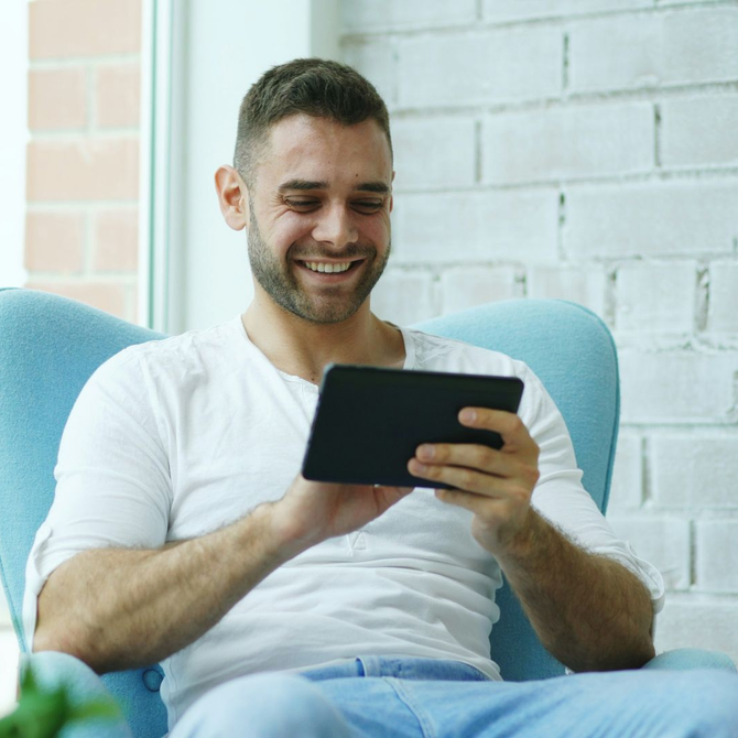 Smiling man in a white shirt using a tablet while sitting in a blue chair