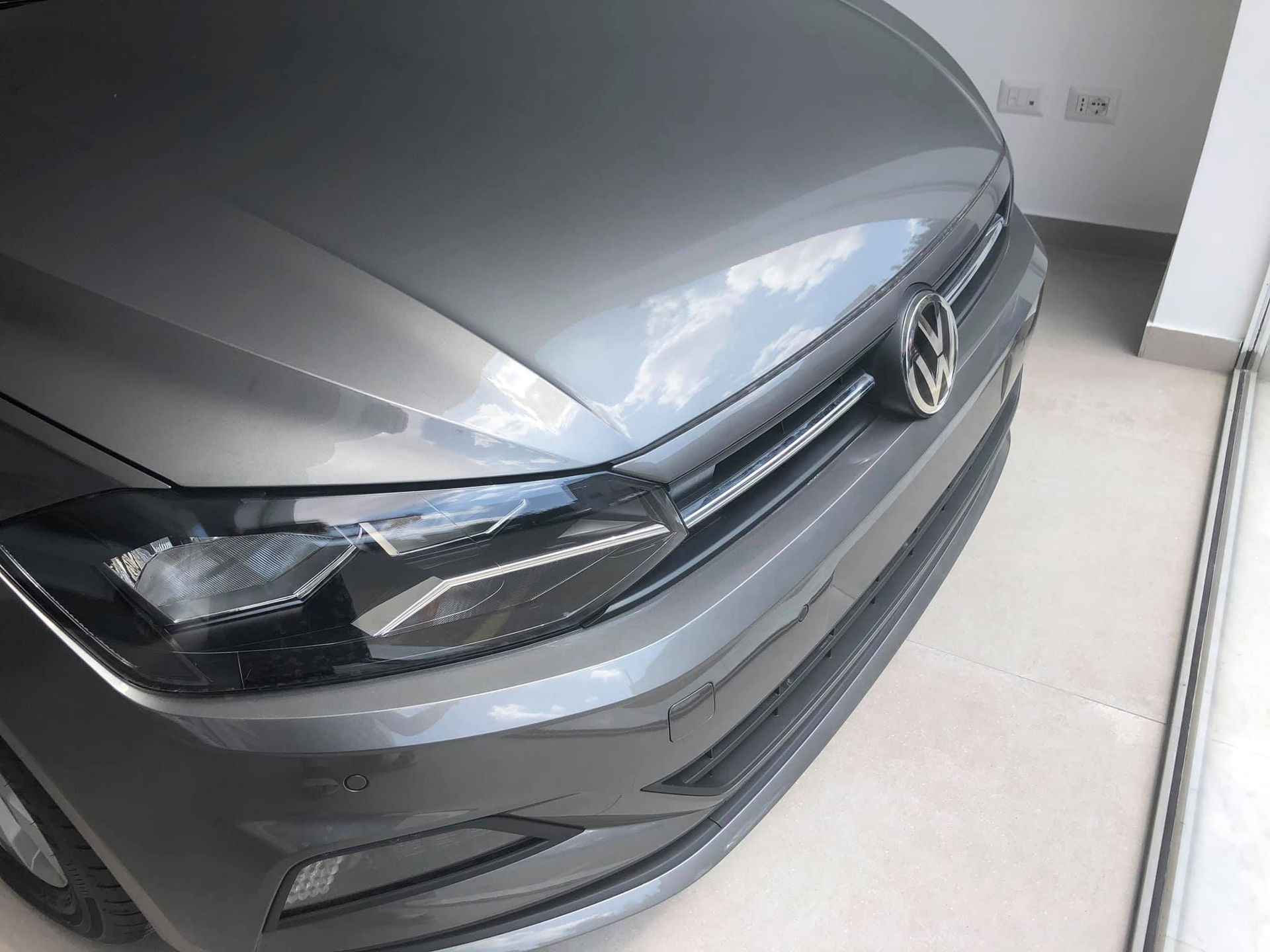 Auto Volkswagen grigia, vista frontale, con fari e stemma visibili, all'interno dello showroom Autoquattro.