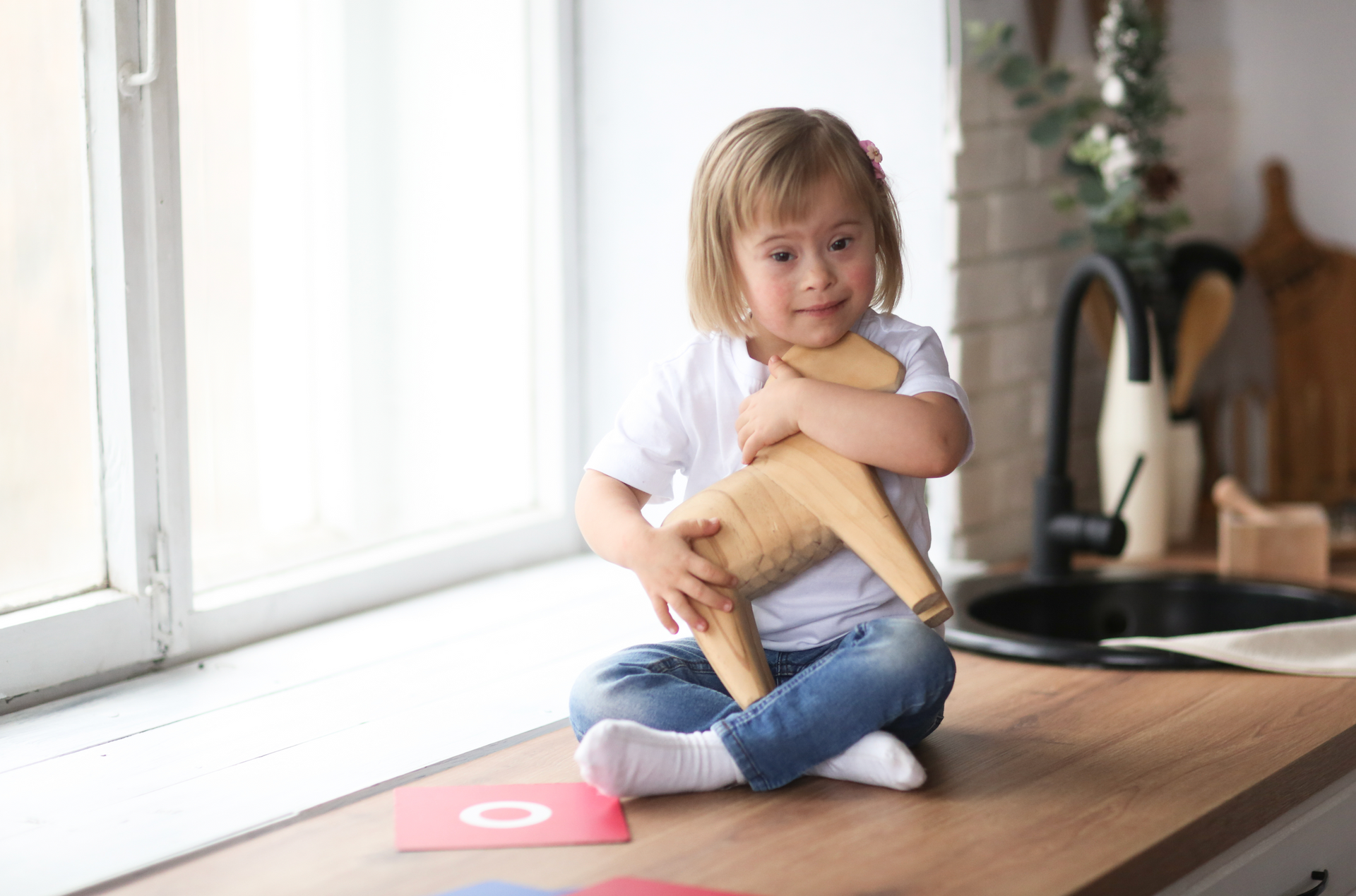 A little girl is sitting on a kitchen counter holding a wooden chair.