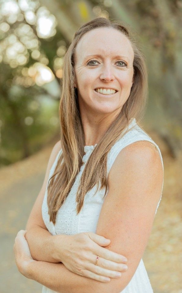 A smiling person with long light brown hair wearing a white sleeveless top, standing outdoors with arms crossed. A smiling person with long light brown hair wearing a white sleeveless top, standing outdoors with arms crossed.