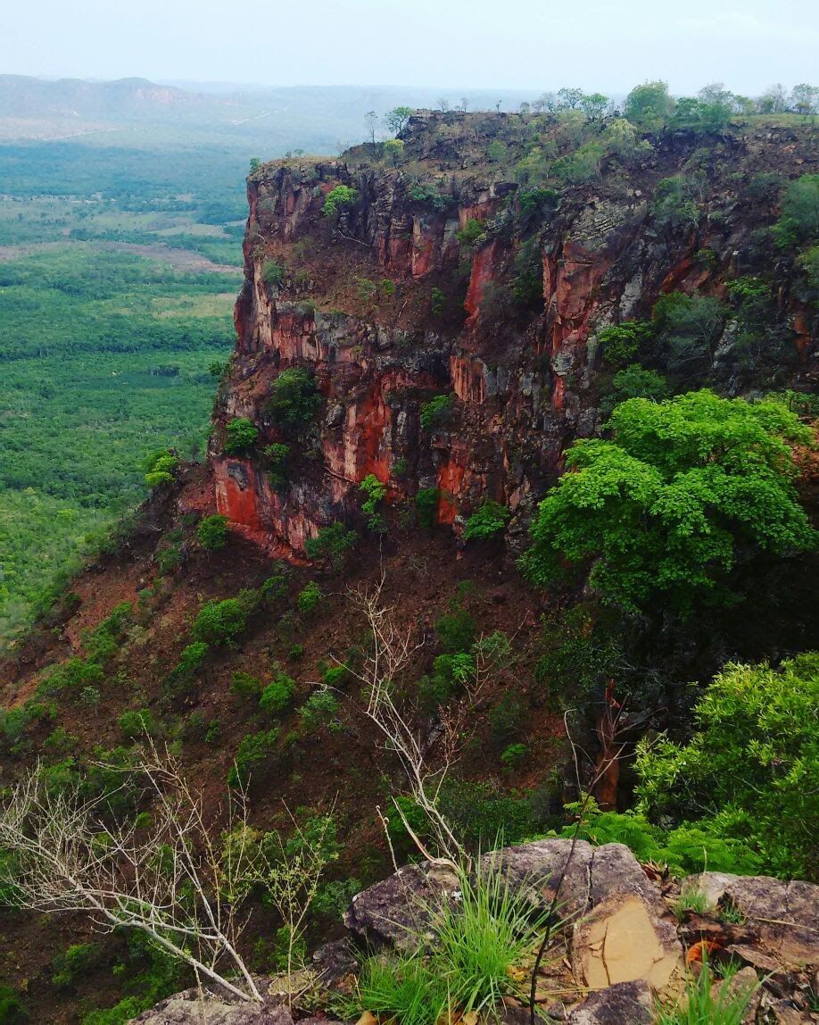 Penhasco rochoso vermelho elevando-se acima de um vale verde sob um céu enevoado.