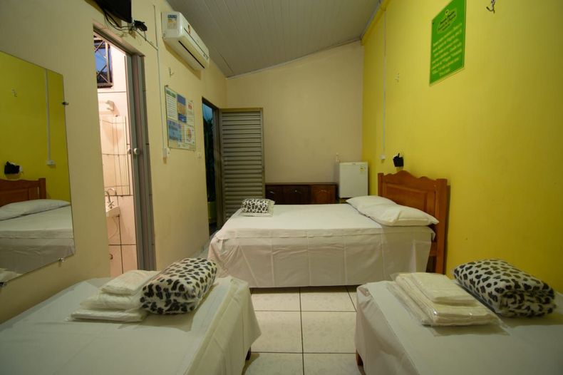 Quarto de hotel pequeno com três camas de solteiro, paredes amarelas e uma porta que dá acesso ao banheiro.