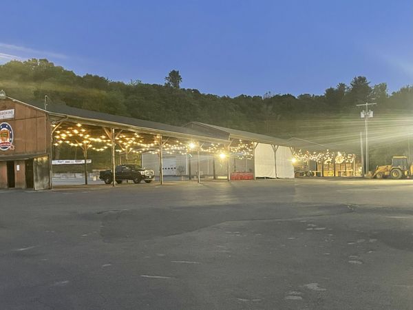 A dark parking lot with buildings, strings of lights, and a forested hillside at dusk.