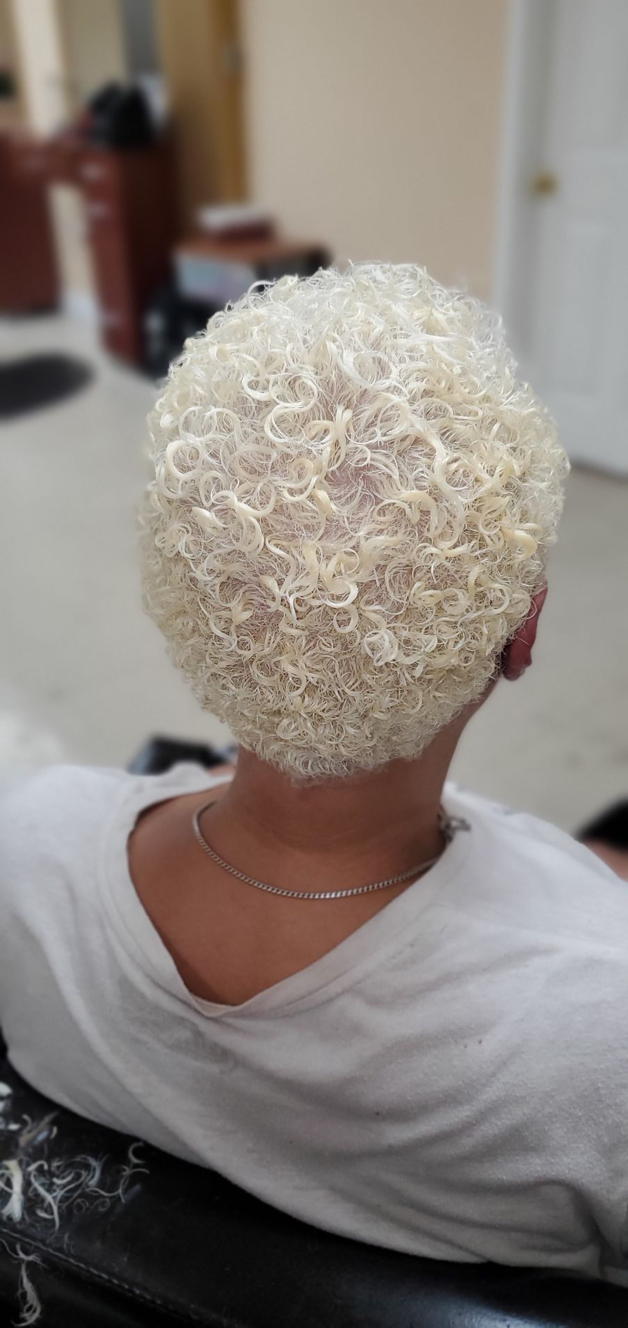 Person seen from behind with short, tightly curled platinum-blond hair, seated indoors in a white shirt.
