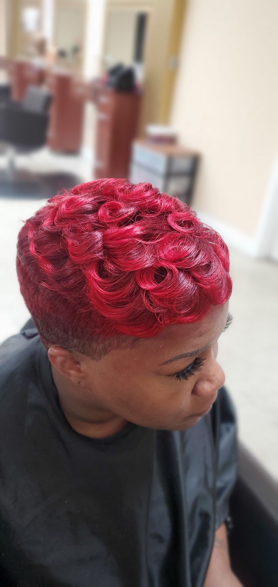 Person with bright red curly hairstyle sitting in a salon chair, viewed from the side.