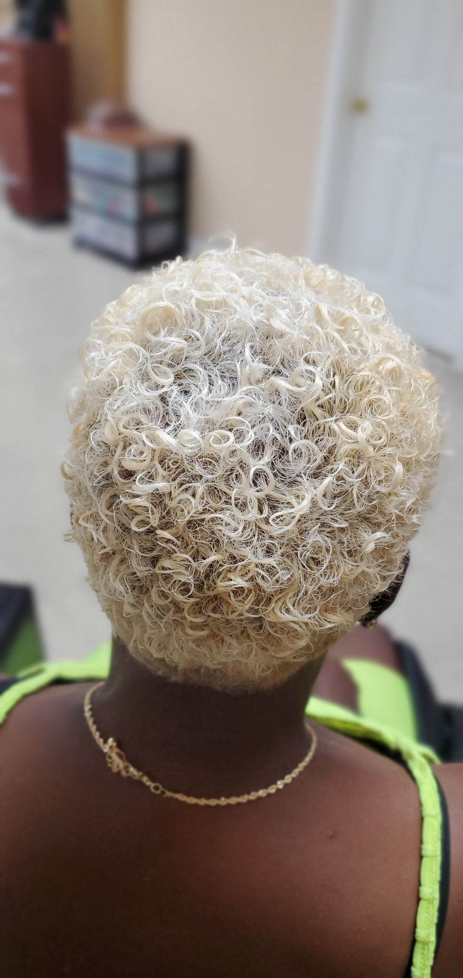 Back view of a person with short blonde curly hair in a dark top and green straps indoors