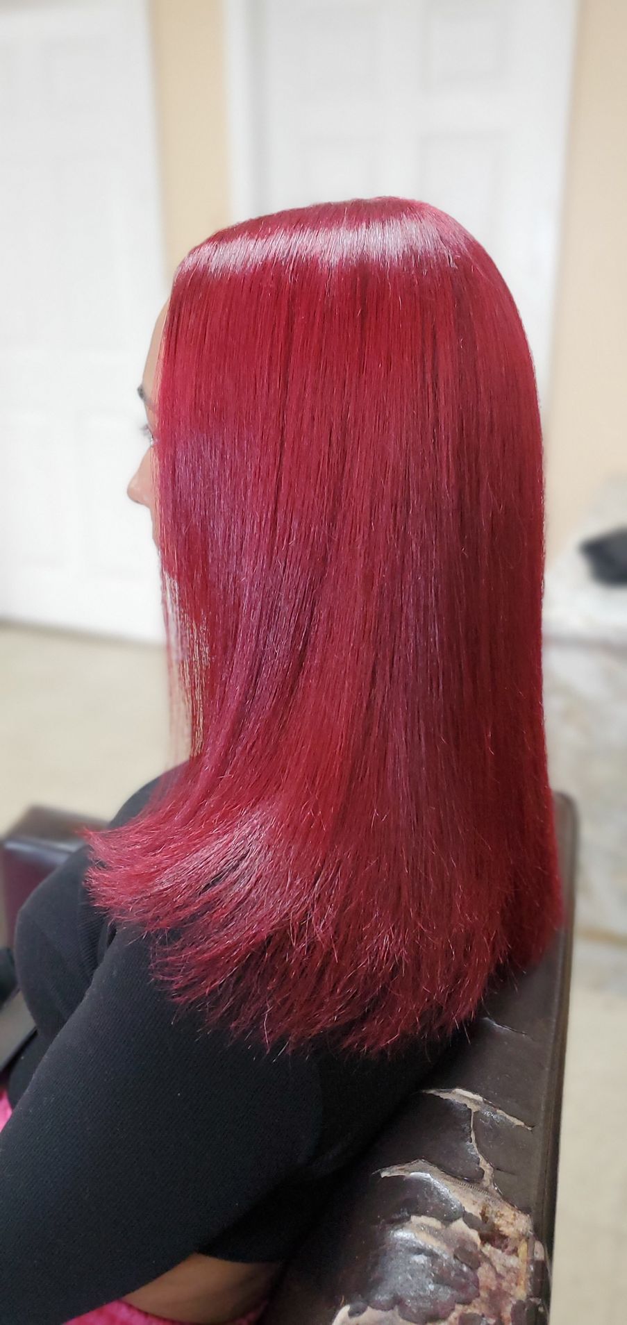 Person with long straight bright red hair seated in a salon chair, viewed from the side.