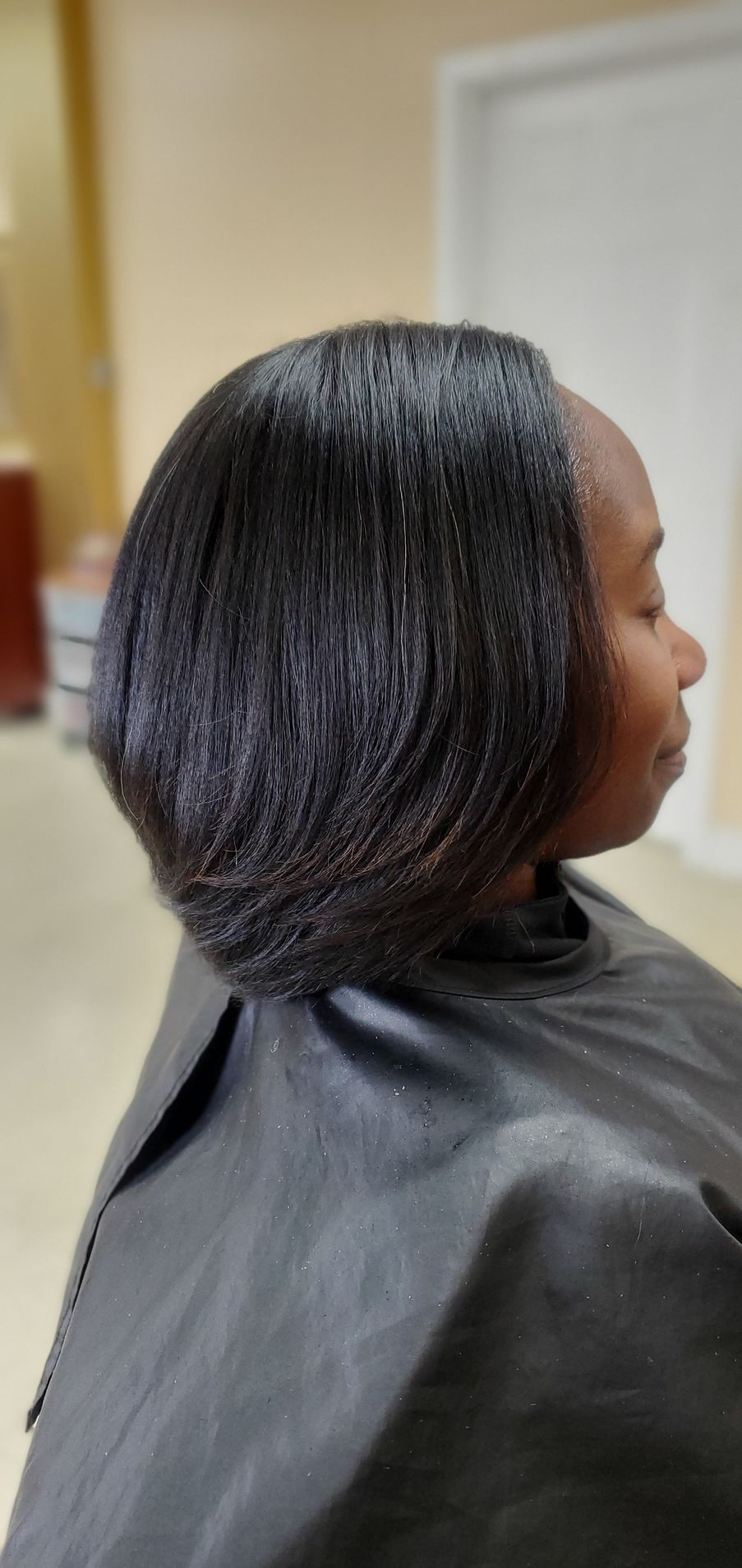 Woman with sleek shoulder-length black hair in a salon chair, seen in profile wearing a black cape.
