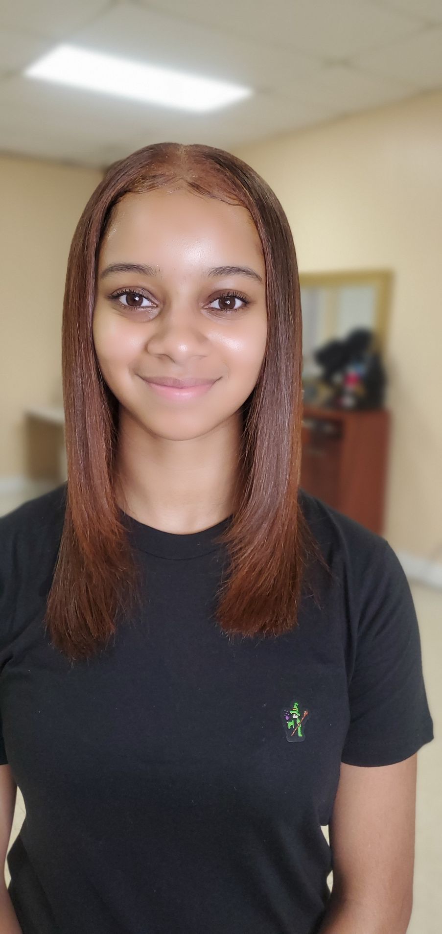 Smiling person with straight braided hair in a black shirt, standing indoors against a softly blurred background