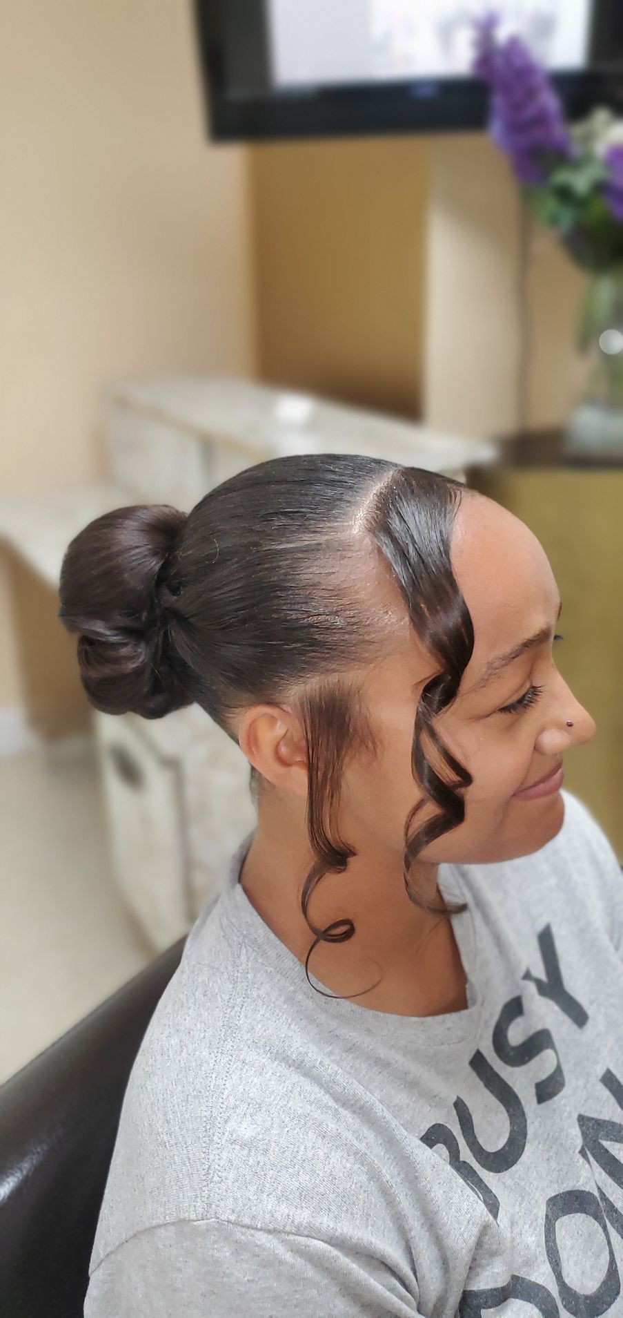 Side profile of a woman with a sleek bun and curled face-framing strands, wearing a gray shirt indoors.